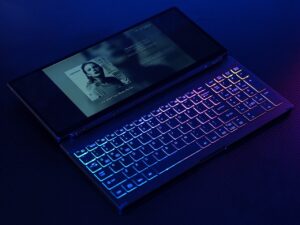A backlit RGB keyboard with a connected touchscreen monitor shows a music player interface. The device is laid out on a dark surface, with the lights glowing in the dark.