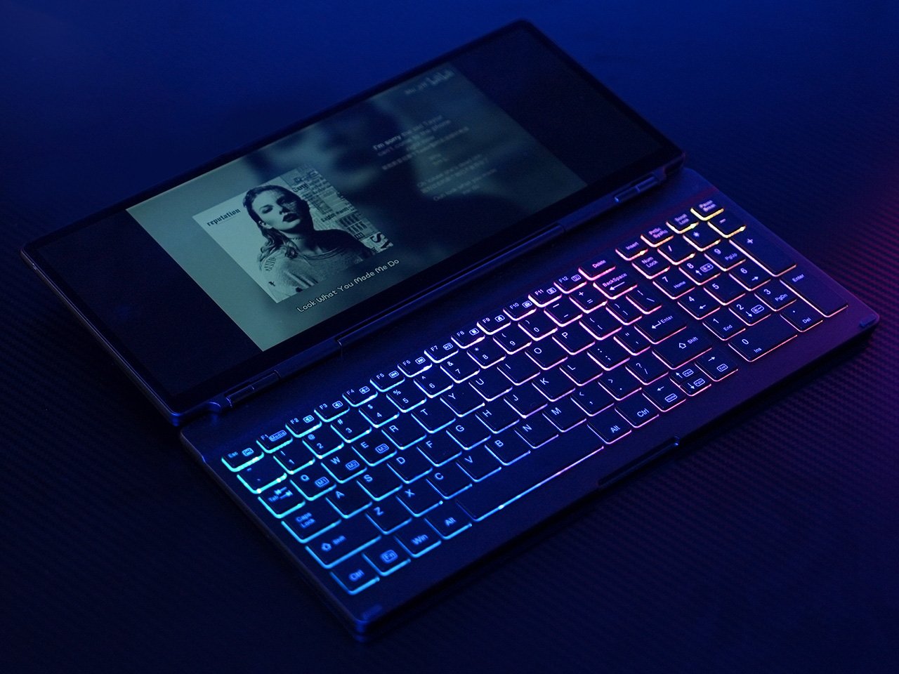 A backlit RGB keyboard with a connected touchscreen monitor shows a music player interface. The device is laid out on a dark surface, with the lights glowing in the dark.